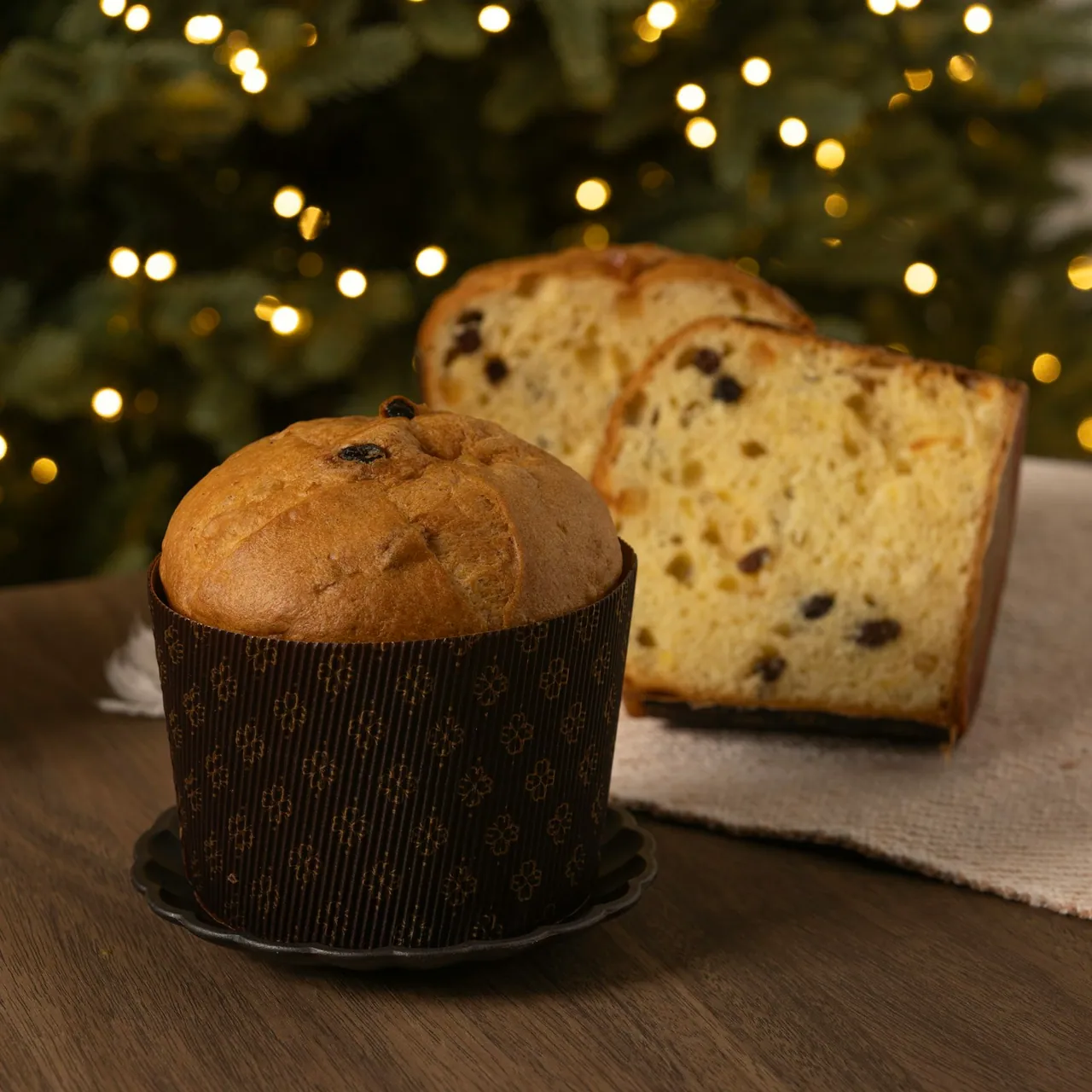 Un panettone entier et un panettone tranché sur une table, devant un sapin illuminé